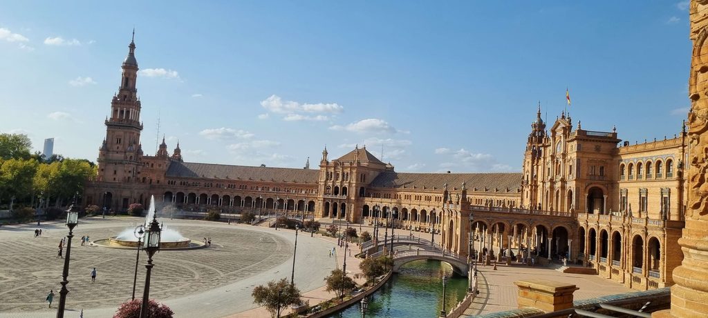 Vue panoramique de la Plaza de España à Séville, avec son bâtiment en brique en arc de cercle, ses tours et son canal, sous un ciel bleu.