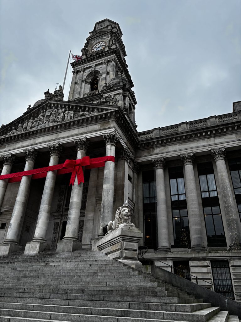 Bâtiment monumental en pierre avec colonnes, horloge et drapeau britannique. Les colones sont entourée d'un imposant ruban rouge pour les fêtes de noël. 