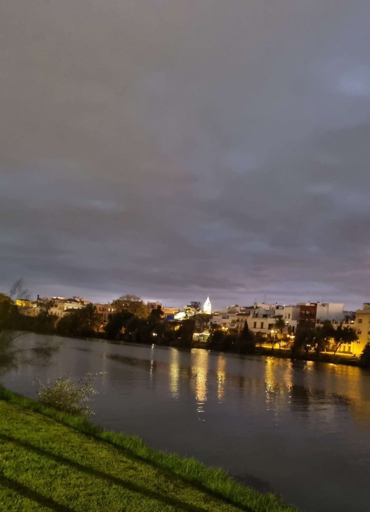 Vue nocturne de Séville depuis les berges du Guadalquivir, avec les lumières de la ville se reflétant sur l'eau.