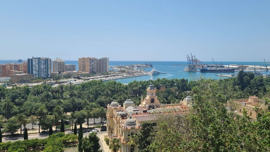 Vue sur le port de Málaga depuis les hauteurs : un paquebot à quai, la mer bleue et, au premier plan, un bâtiment ocre à dômes et à tour d’horloge entouré de palmiers.